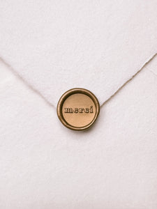 Mini gold wax seal featuring "merci" in a serif font on a white handmade paper envelope