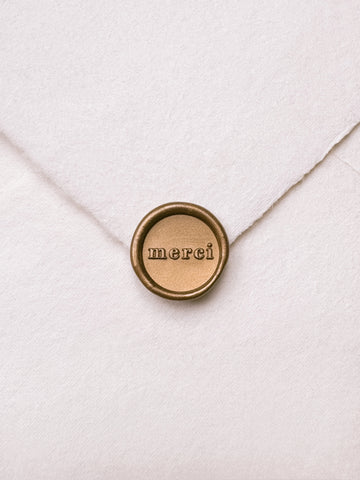 Mini gold wax seal featuring "merci" in a serif font on a white handmade paper envelope