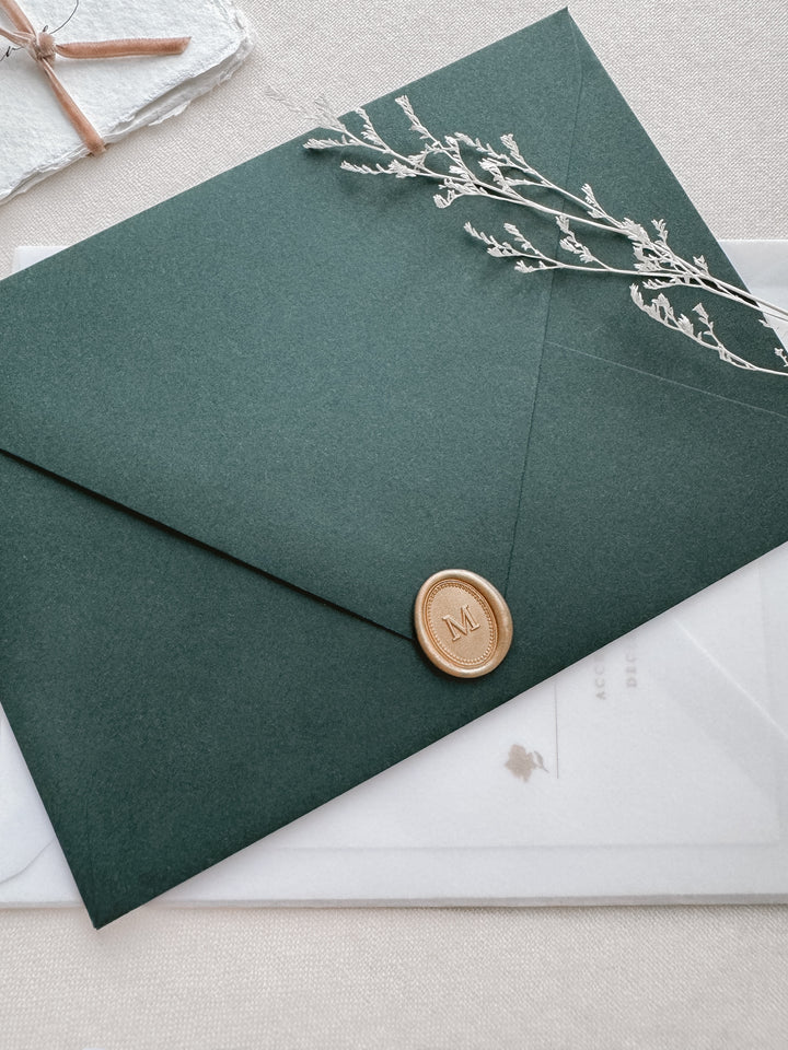 Personalized initial mini gold wax seal featuring a custom monogram framed by an oval line border on a dark green envelope on a wedding invitation vellum envelope