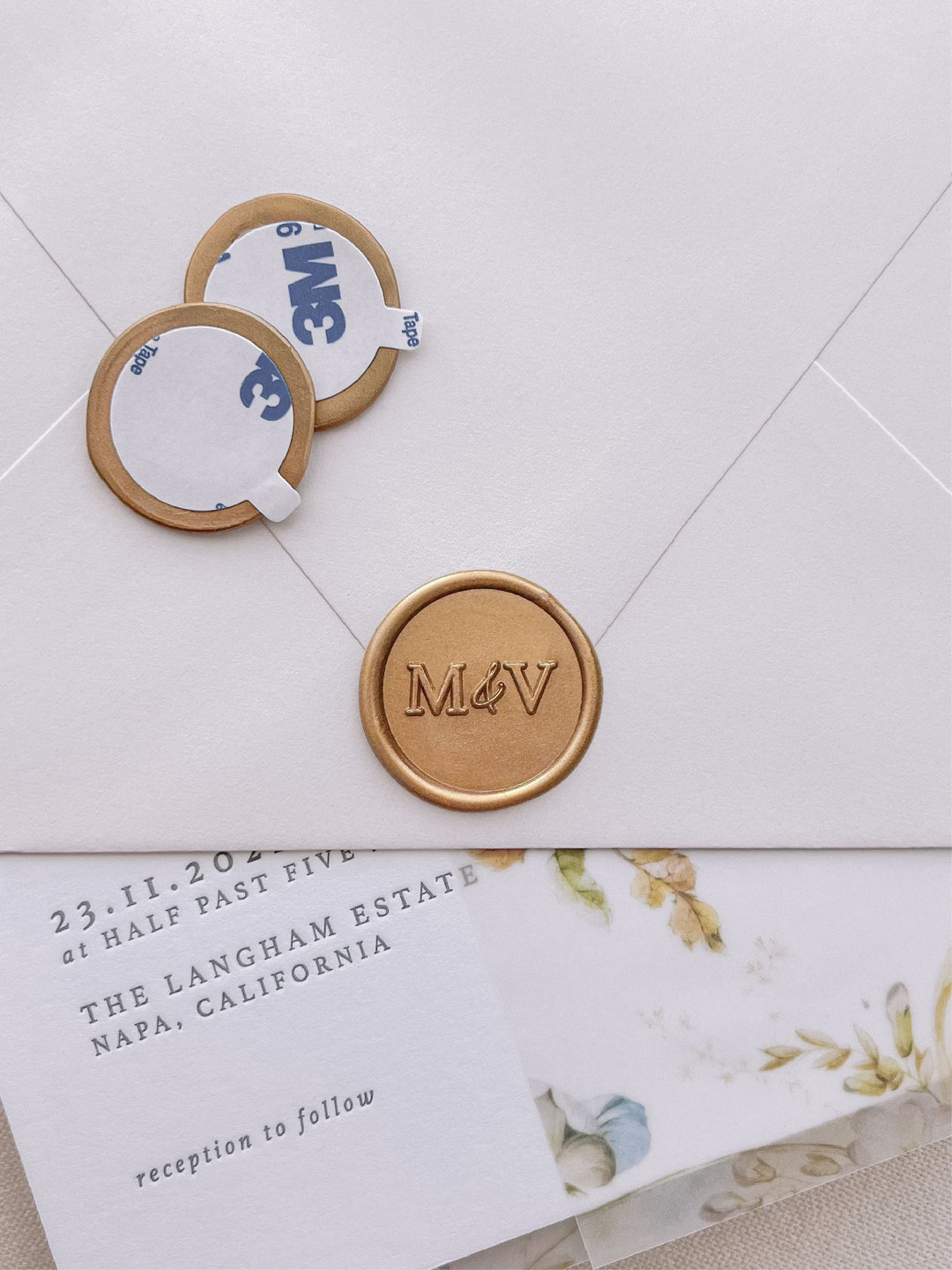 modern monogram with "&" gold wax seal stickers with self-adhesive backings on a wedding invitation, beige envelope, and floral vellum jacket