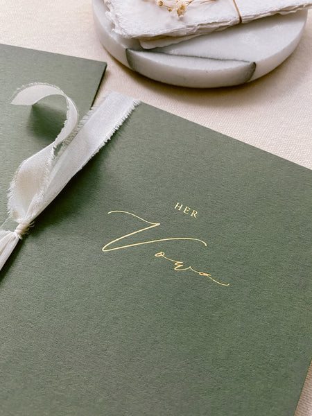 A set of two His and Her gold foil olive green card stock vow books in calligraphy script with antique white colored silk ribbon