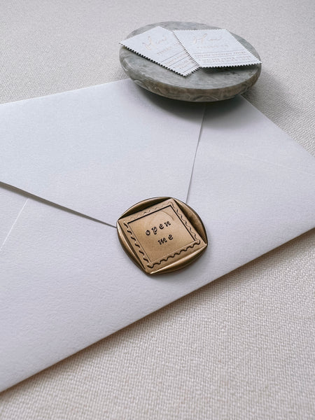 Open me wax seal in gold on paper envelope styled with small gray stone dish with Olive Paperie Co logo stamps