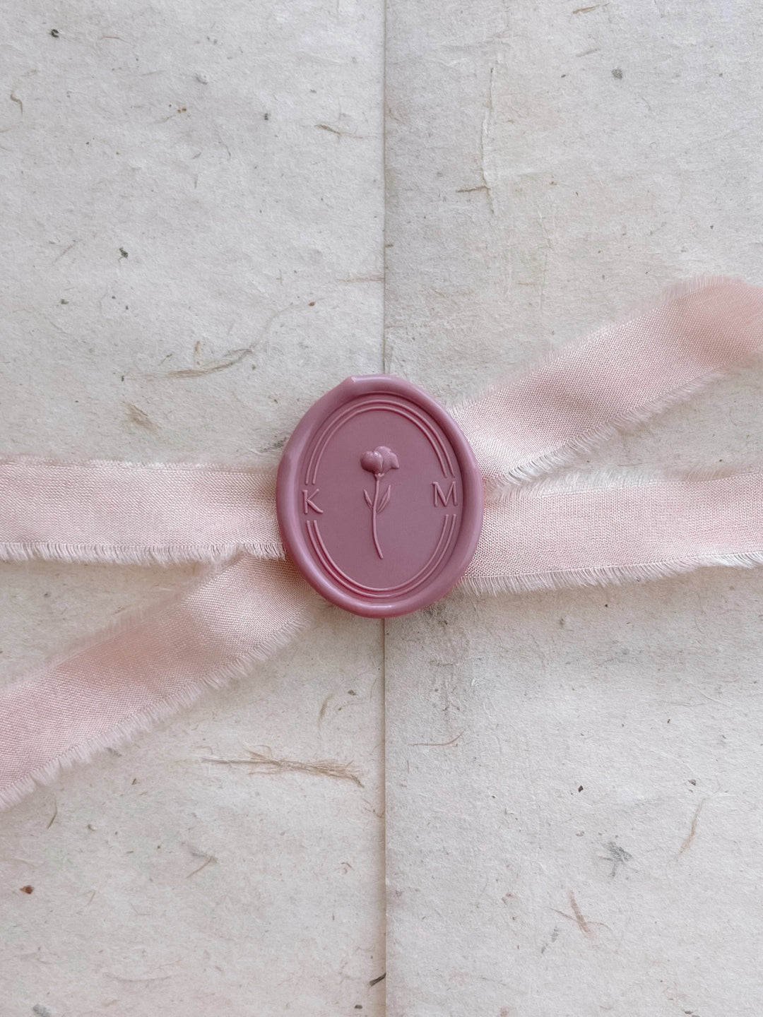 Dusty pink personalized initials 3D floral oval wax seal featuring a custom monogram on a light pink silk ribbon tied on a light beige textured envelope wrap