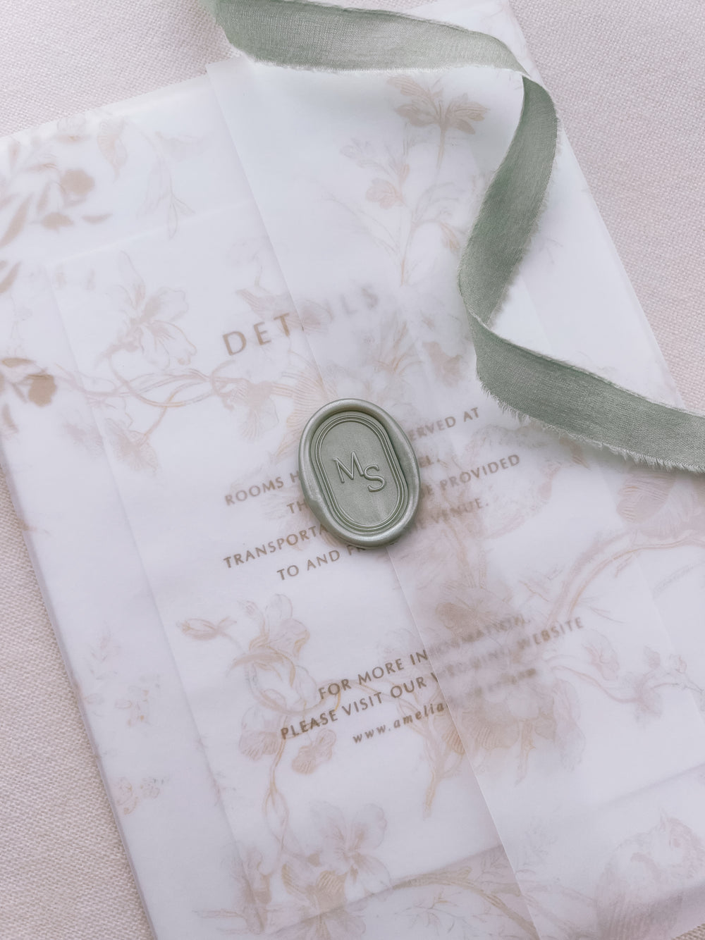 Light metallic sage green personalized initials oval arch wax seal featuring a custom monogram framed by simple double line border on a beige floral wedding invitation vellum jacket