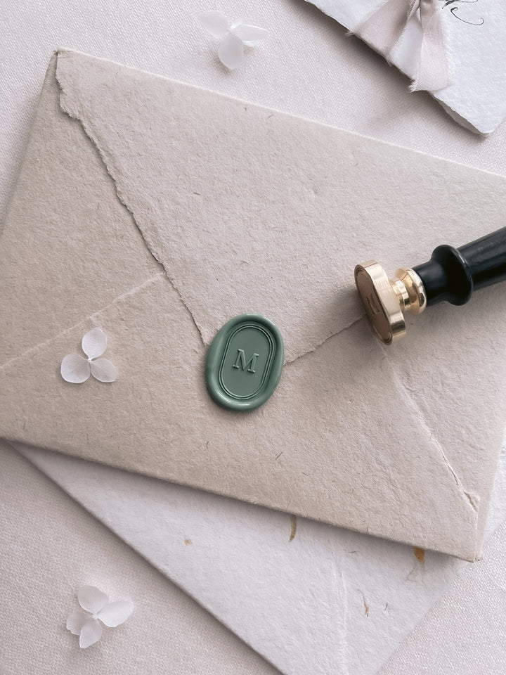 Sage green personalized single initial oval line border wax seal featuring a custom monogram on a stack of textured envelopes with a wax stamp head and black handle