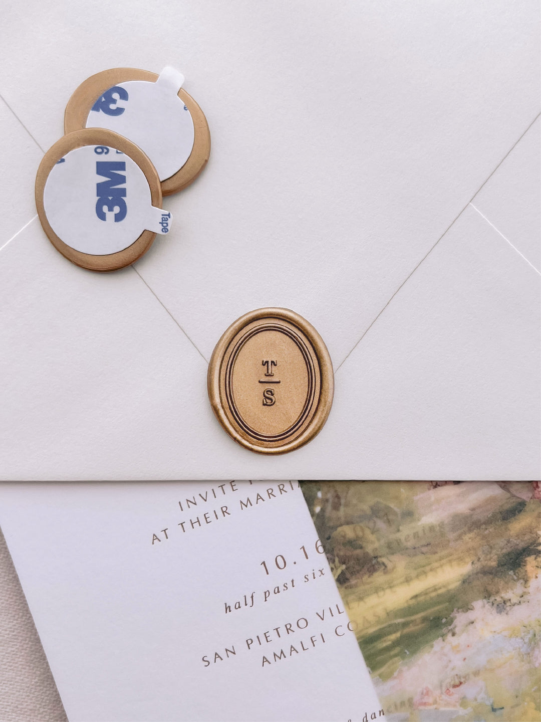 Oval border monogram gold wax seal stickers with self-adhesive backings on a wedding invitation, beige envelope, and vellum jacket
