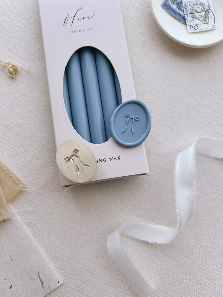 Dusty blue oval wax seal with a bow design, placed on a box of dusty blue sealing wax sticks