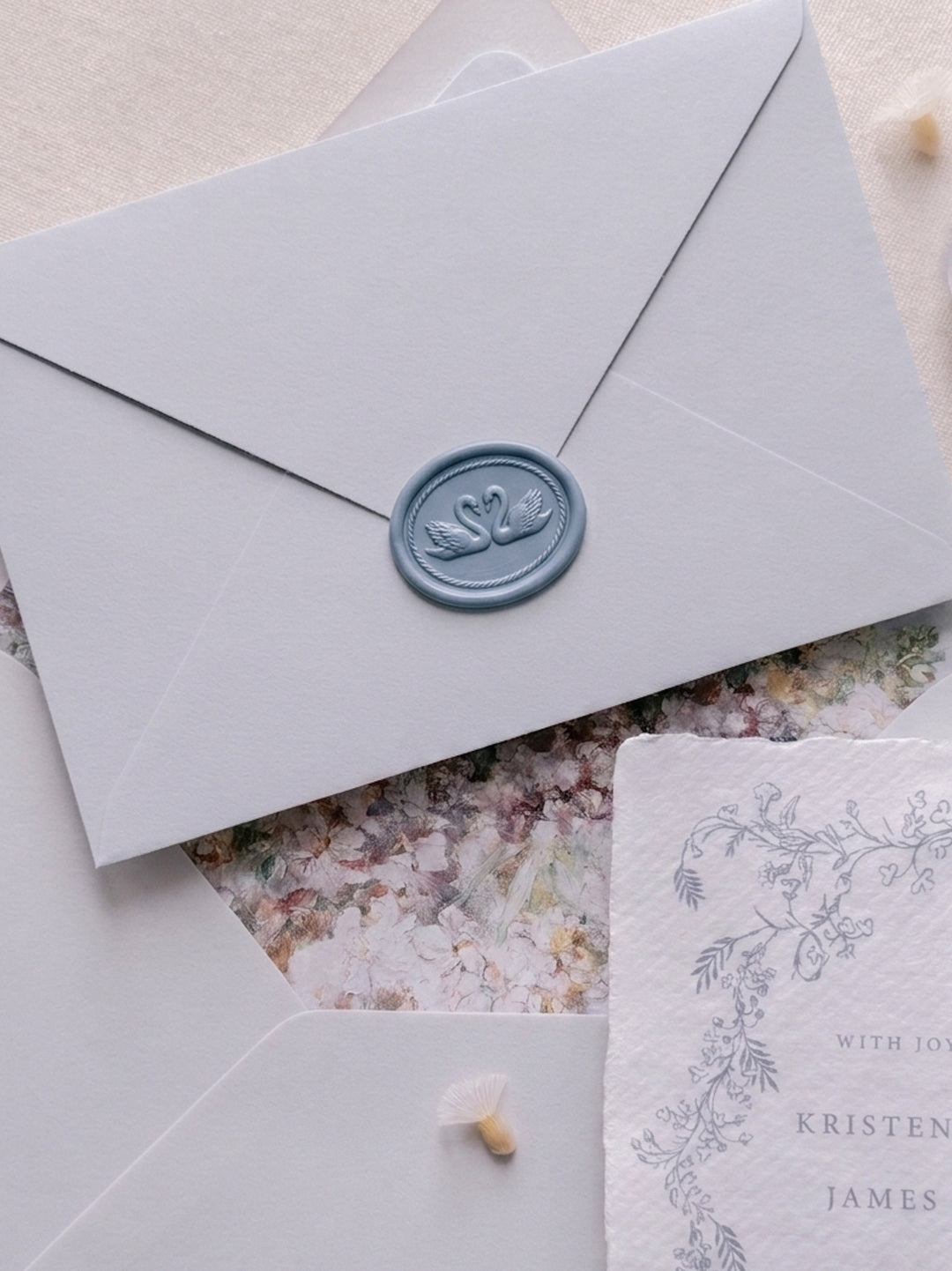 Dusty blue double swan oval wax seal on a light blue envelope over a blue botanical wedding invitation card and an envelope with a floral envelope liner