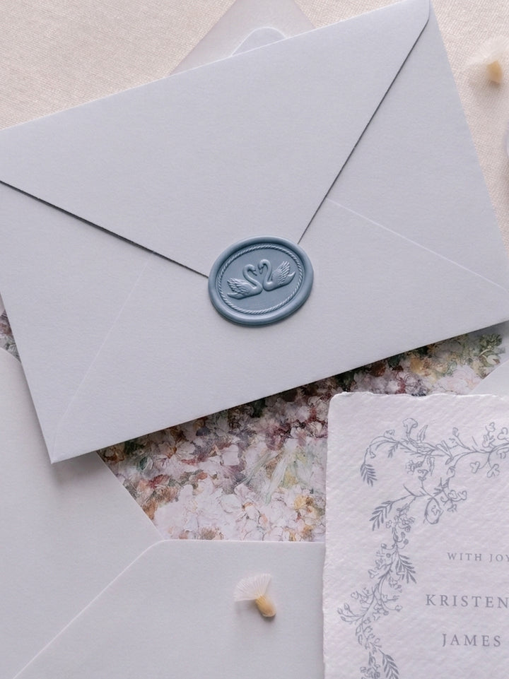 Slate blue double swan oval wax seal on a light blue gray wedding invitation envelope, layered over a floral-lined envelope and botanical wedding invitation suite.