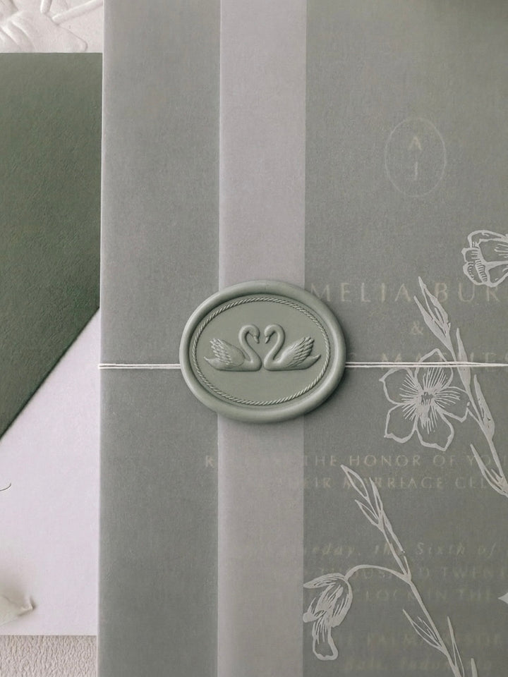 Light sage green oval wax seal featuring a design of two swans framed by a rope style decorative oval shaped border, on a floral vellum jacket over an olive green wedding invitation suite