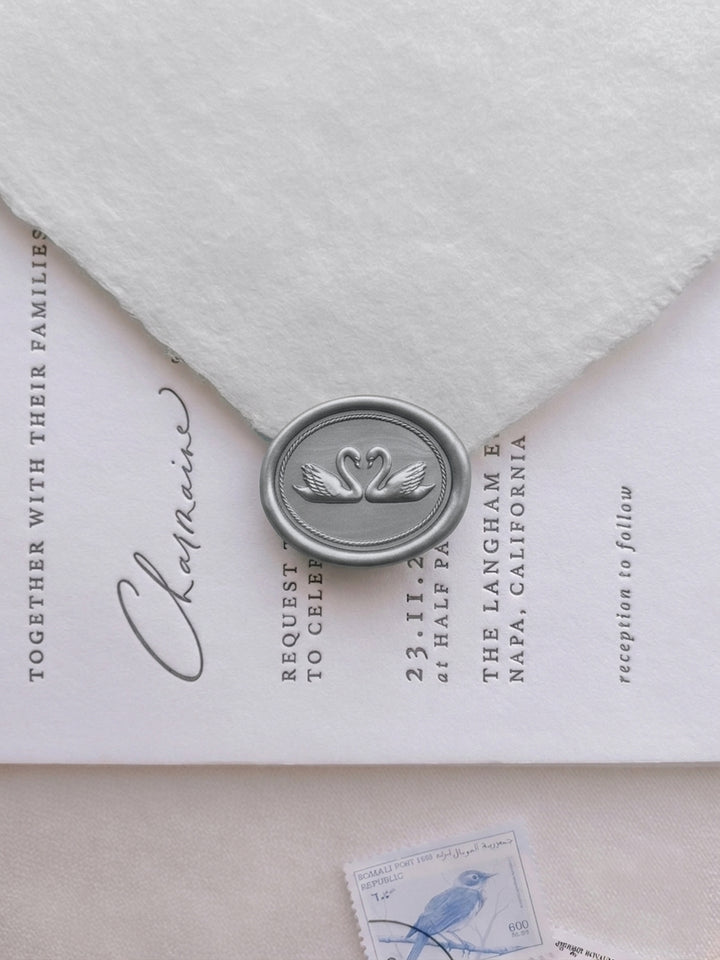 Silver double swan oval wax seal on a deckle-edge white wedding envelope, set over a modern calligraphy wedding invitation with vintage postage stamp detail.
