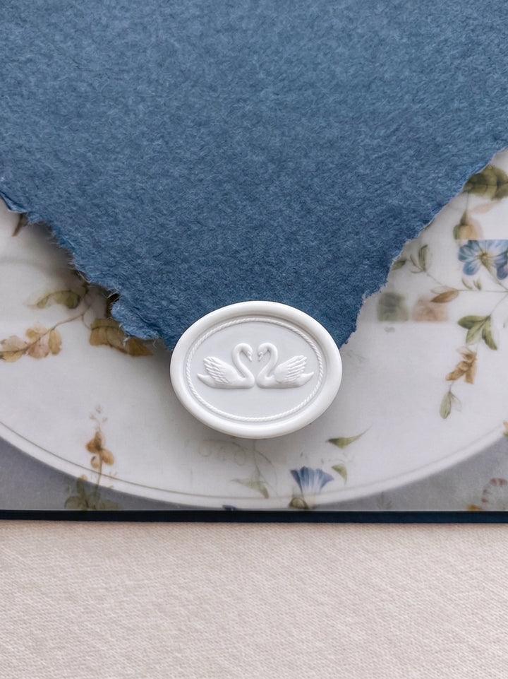 White double swans oval wax seal on a blue deckled edge handmade paper envelope flap over a floral vellum jacket and wedding invitation card