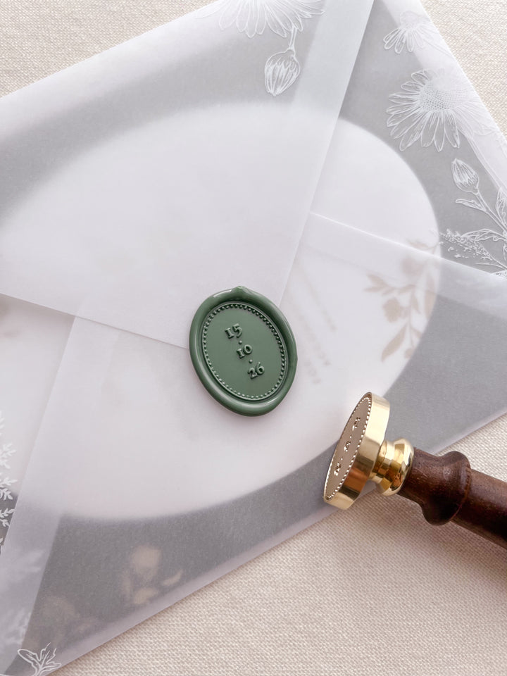 Sage green custom date oval wax seal personalized with a wedding date framed by a dotted border on a wedding invitation vellum jacket with a wax stamp head and wooden handle