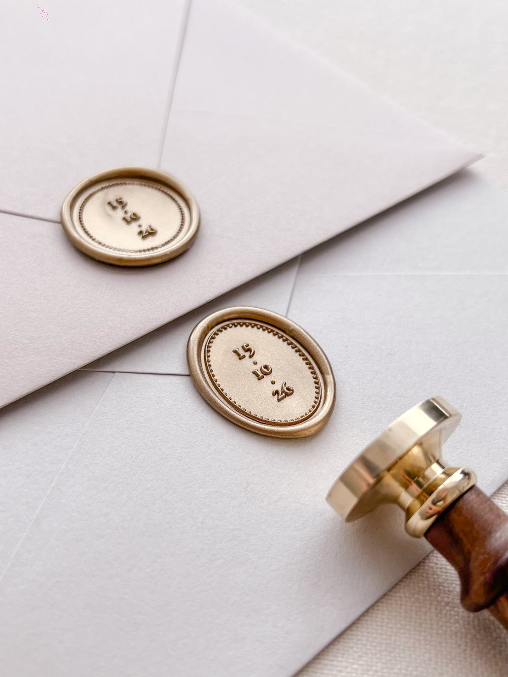 Gold personalized wax seals featuring custom wedding dates on light grey envelopes with a wax stamp head and a wooden handle