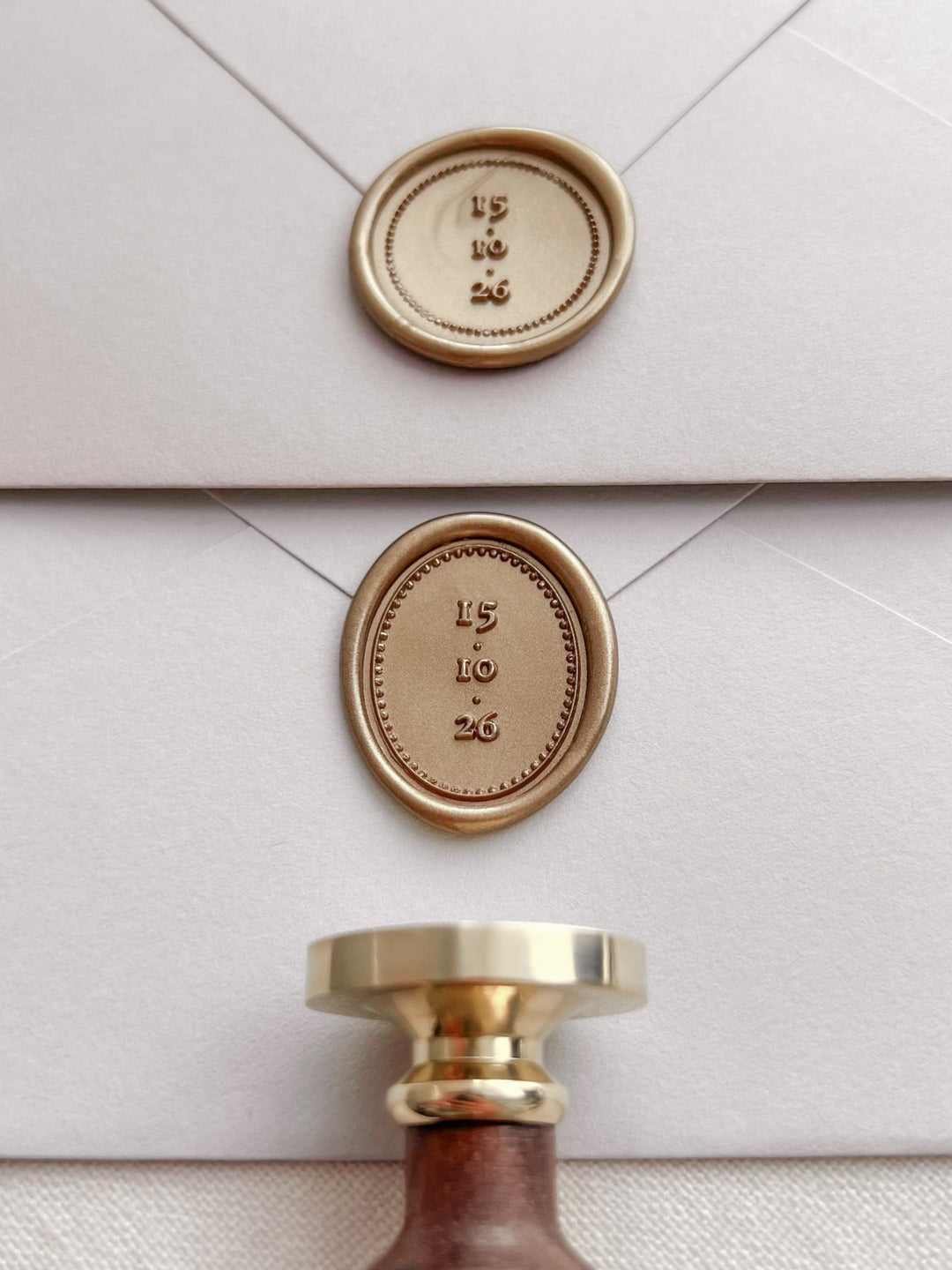Personalized gold oval and round wax seals featuring custom wedding dates on light grey envelopes with a wax stamp head and wooden handle 