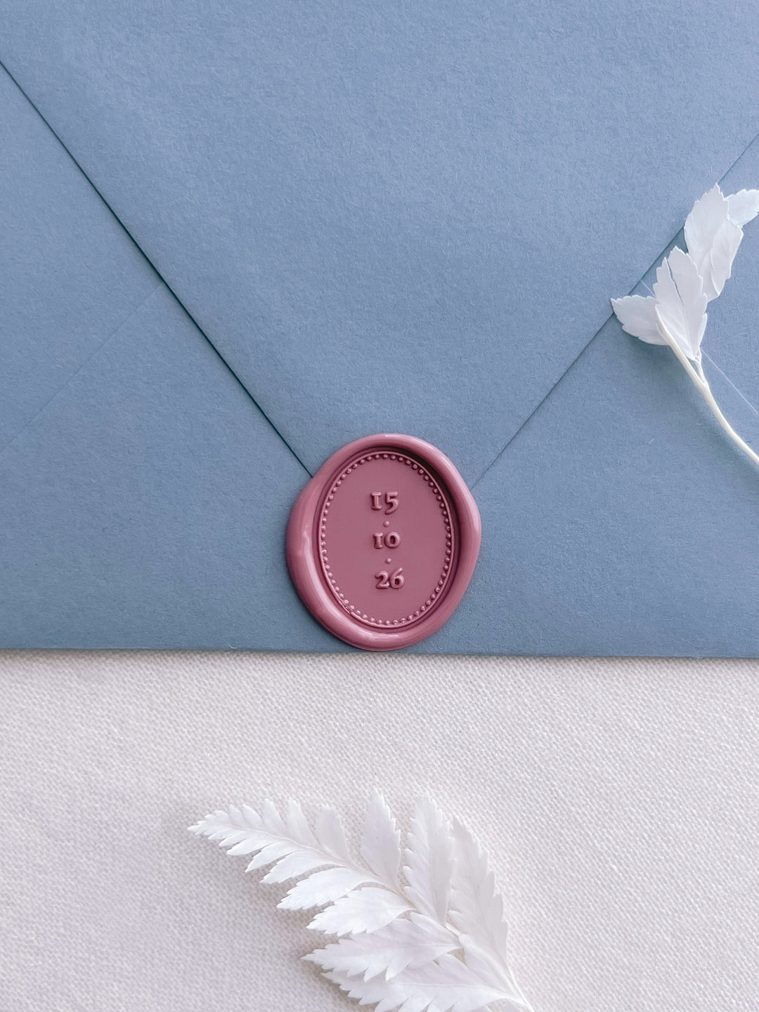 Rose pink personalized oval wax seal featuring a custom wedding date and an oval border on a dusty blue envelope on a white background