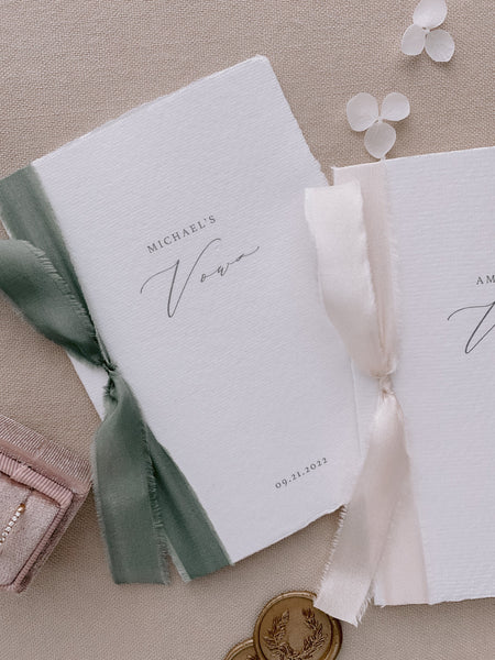 A set of two His and Her personalised handmade paper wedding vow books in calligraphy script with one tied with nude colored silk ribbon and the other tied with olive colored silk ribbon styled with bride wedding rings and gold wax seals