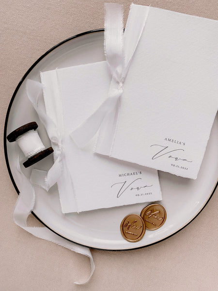 A set of two His and Her personalised handmade paper vow books in calligraphy script with soft white colored silk ribbon styled with gold wax seals and spool of soft white silk ribbon