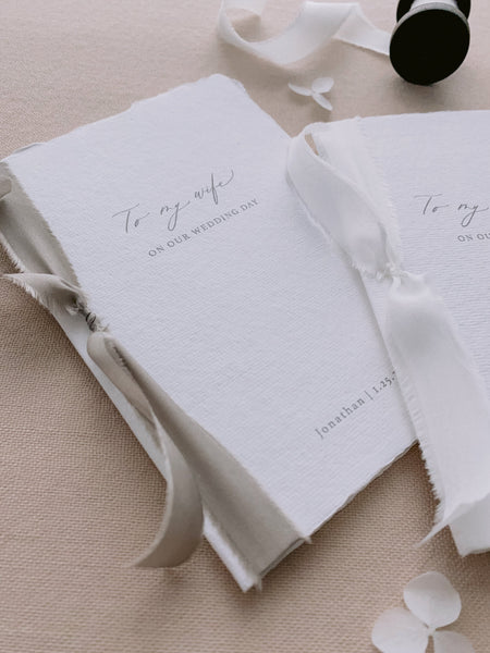 A set of two His and Her personalised wedding love letters handmade paper vow books in modern calligraphy script with one tied in soft white colored silk ribbon and the other tied with sage colored silk ribbon styled with a spool of soft white silk ribbon