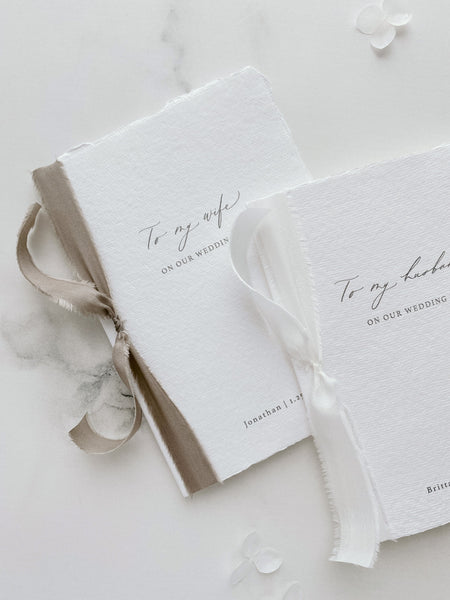 A set of two His and Her personalised wedding love letters handmade paper vow books in modern calligraphy script with one tied in soft white colored silk ribbon and the other tied with sage colored silk ribbon styled with flower petals