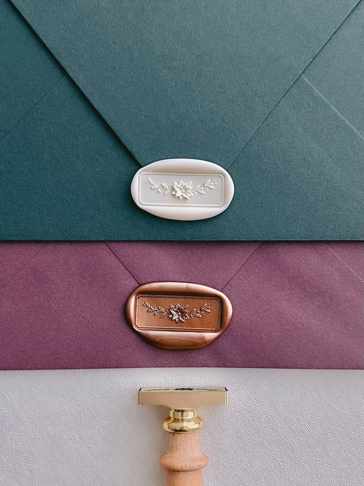 Off-white and antique bronze rectangular Christmas Poinsettia garland wax seals on a dark teal envelope and a burgundy envelope, with a brass wax stamp head and wooden at the bottom 