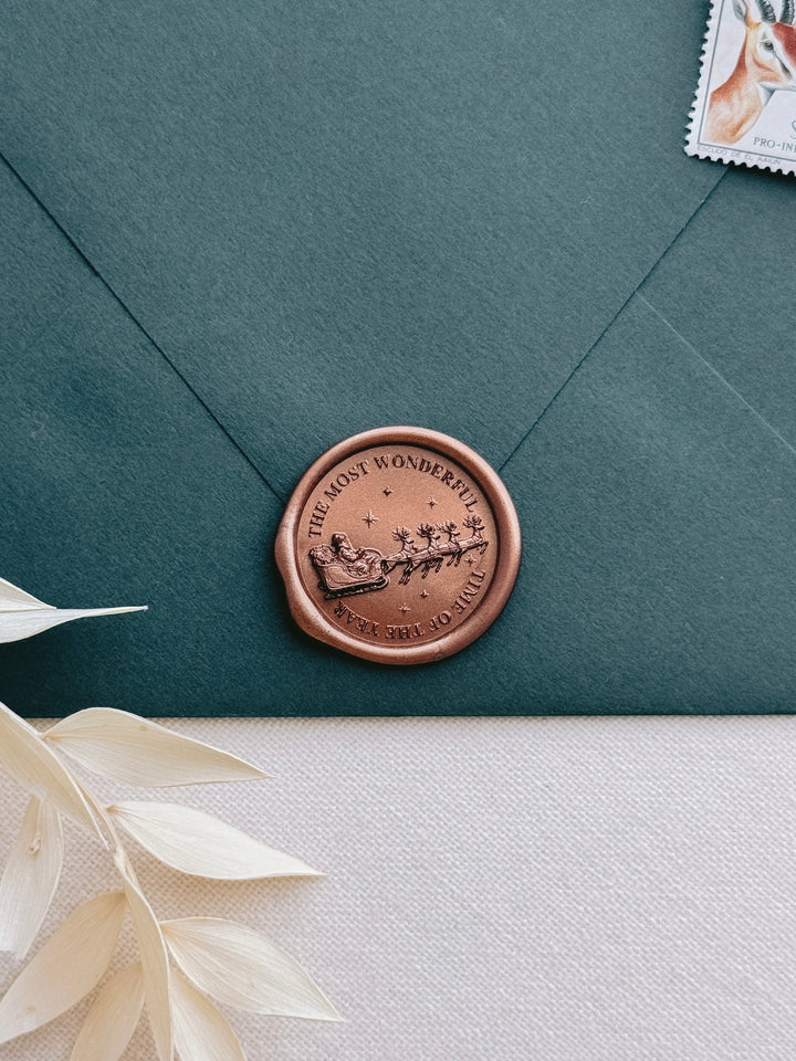 A dark teal-green envelope sealed with a round copper-toned wax seal featuring an engraved Santa Claus and deer sleigh and the text &
