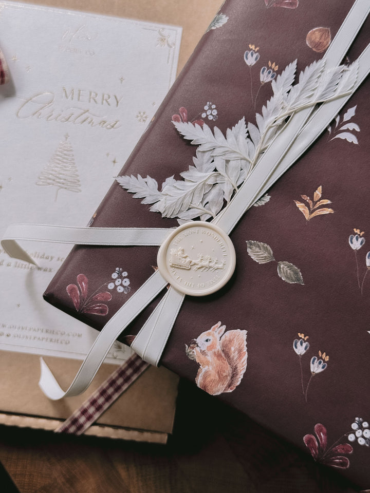 Off-white Santa sleigh wax seal on festive burgundy woodland gift wrap, tied with ivory ribbon and dried foliage.