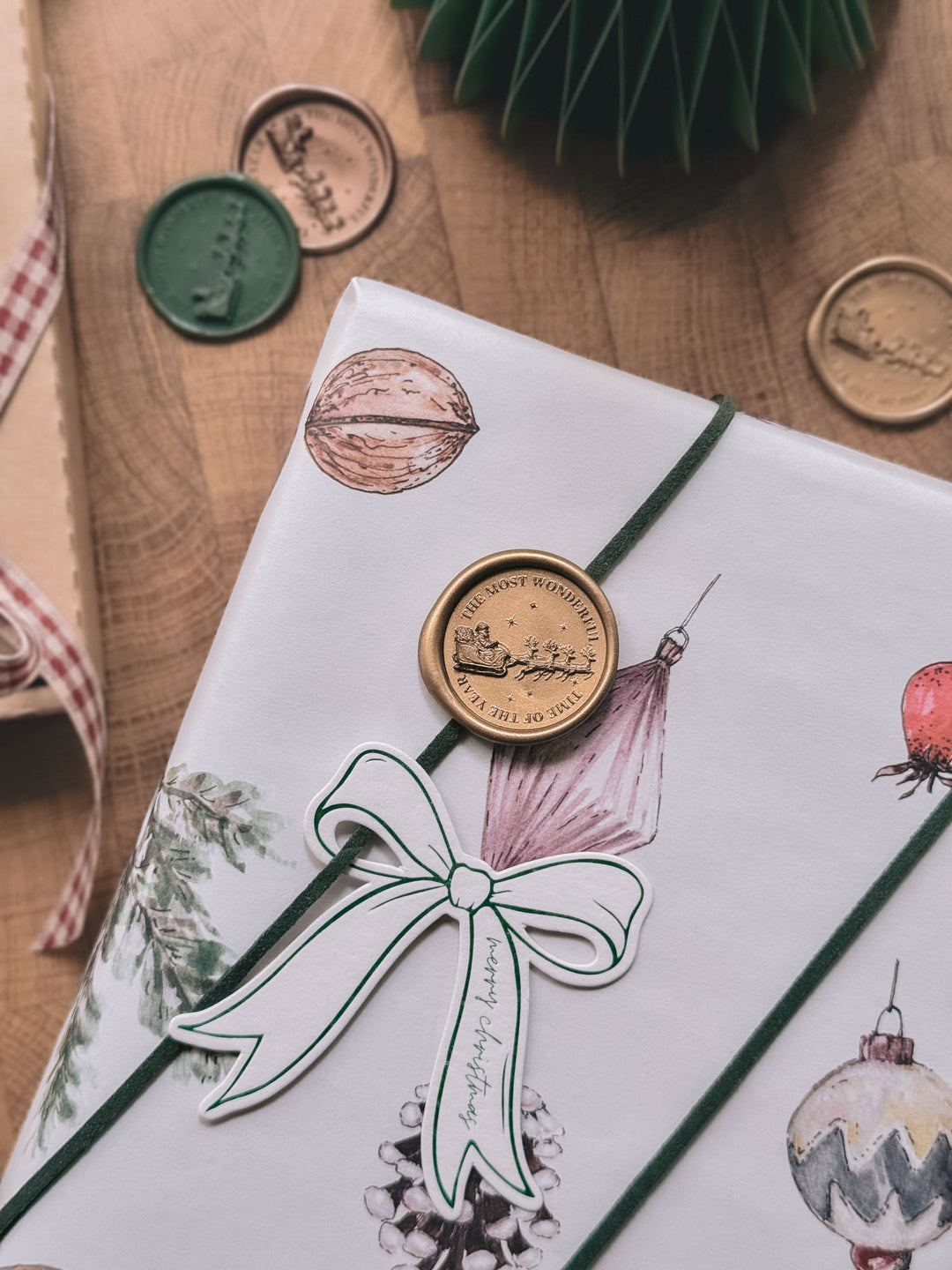 Gold Santa sleigh wax seal on white ornament-print gift wrap with a bow-shaped gift tag, alongside gold and green wax seals on a wooden surface.