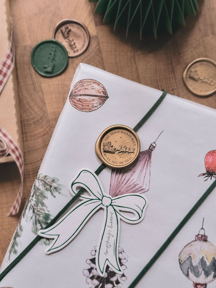 Gold Santa sleigh wax seal on white ornament-print gift wrap with a bow-shaped gift tag, alongside gold and green wax seals on a wooden surface.