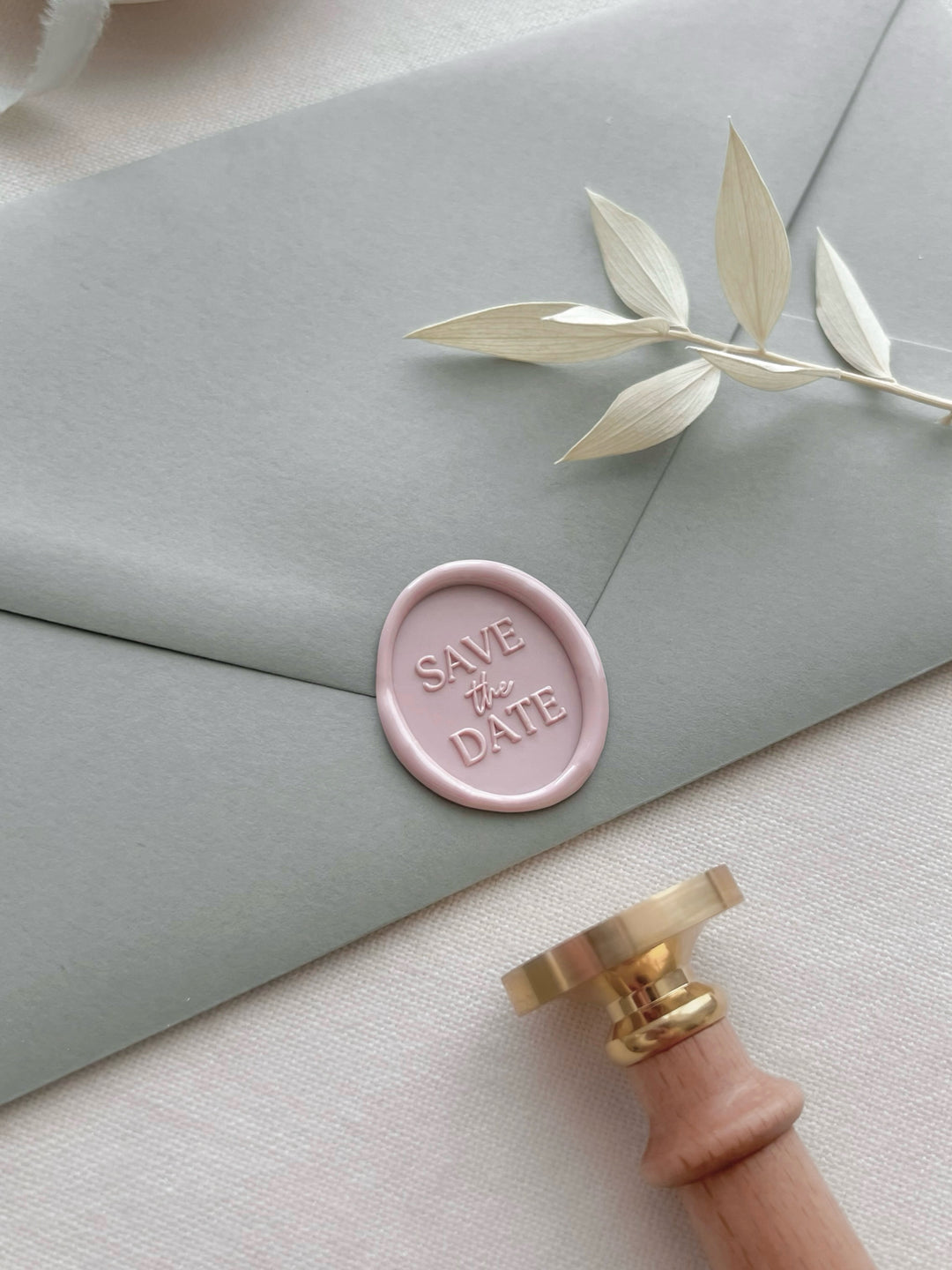 Pink save the date oval wax seal on a light green envelope with a wax stamp head and light wooden handle