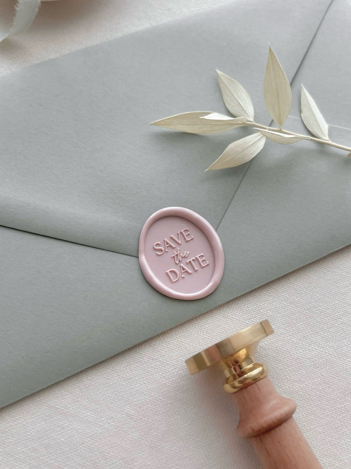 Pink save the date oval wax seal on a light green envelope with a wax stamp head and light wooden handle