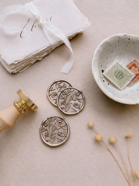 White gold floral wax seals styled with handmade paper place cards, a wax seal stamp, and a small dish with postage stamps