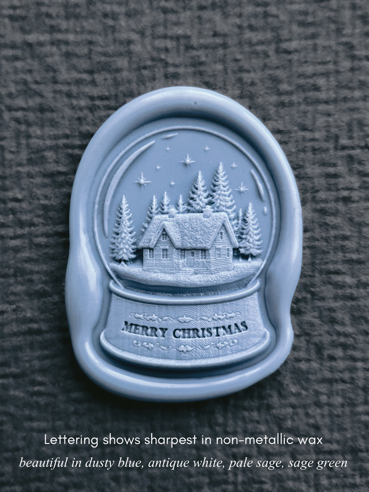 close up shot of a dusty blue snow globe wax seal in a die-cut shape featuring a snow dusted cabin in a forest under a starry sky, with &