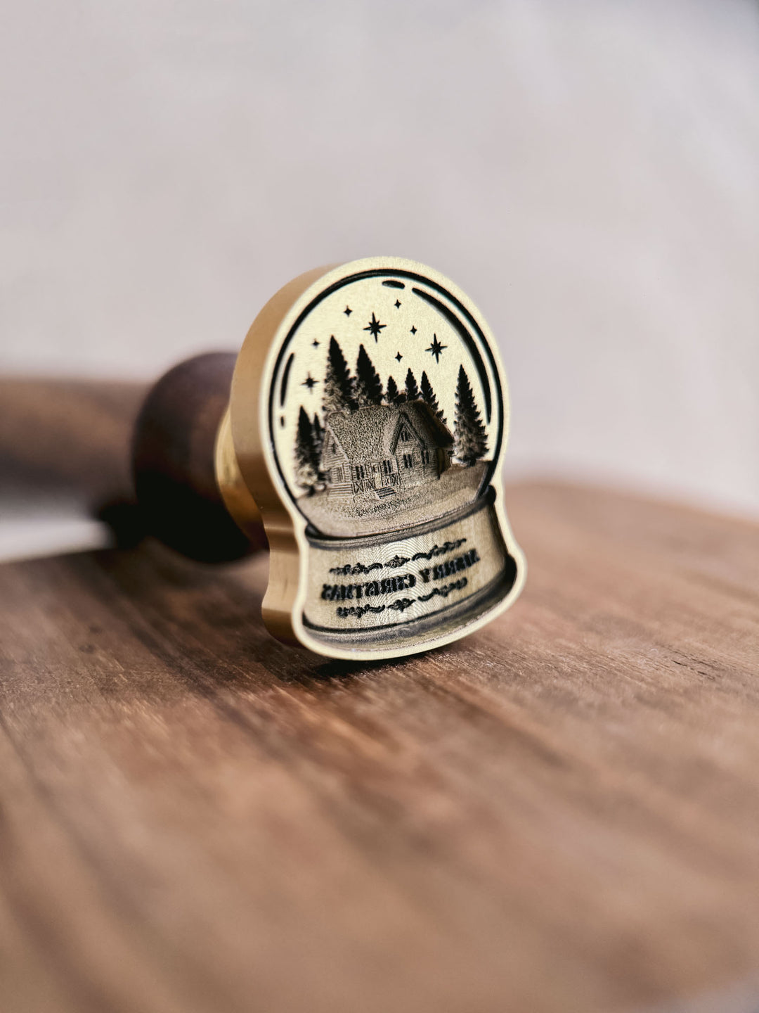 Merry Christmas snow globe wax seal stamp, showing the die-cut brass stamp head engraved with a cabin, pine trees and lettering, resting on a wooden surface.