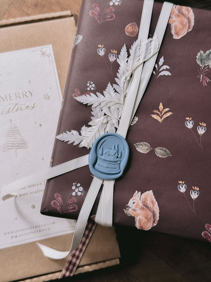 Dusty blue die-cut Merry Christmas snow globe wax seal on festive burgundy botanical gift wrap, tied with ivory ribbon and dried foliage beside a Christmas gift.