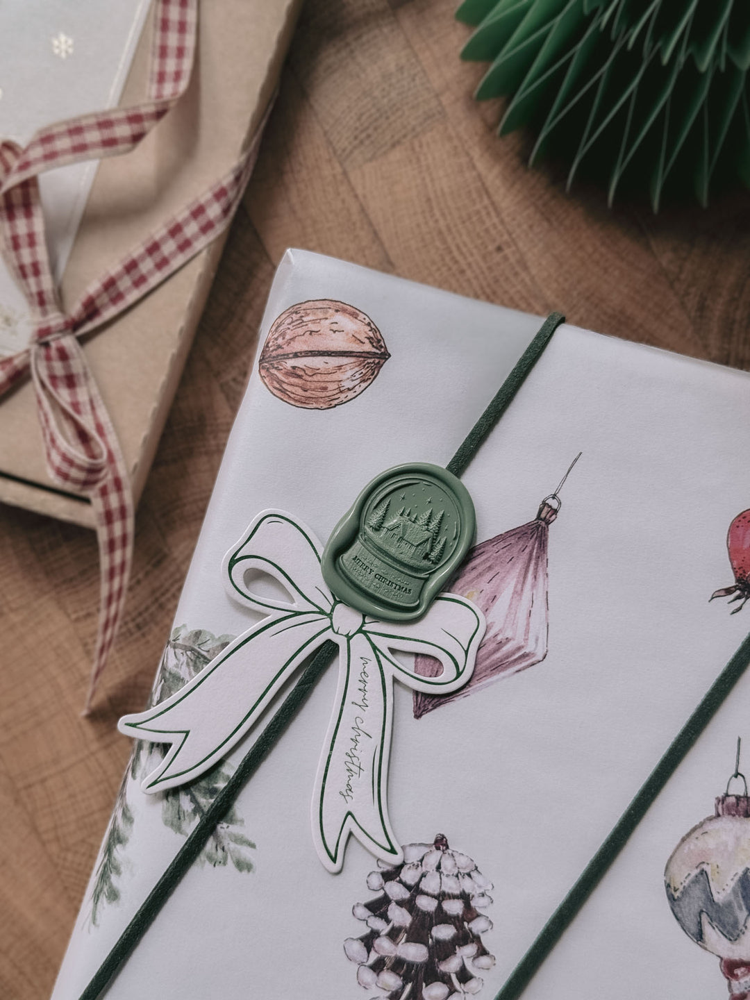 Sage green die-cut Merry Christmas snow globe wax seal on white ornament-print gift wrap, paired with a bow-shaped gift tag and green suede twine.