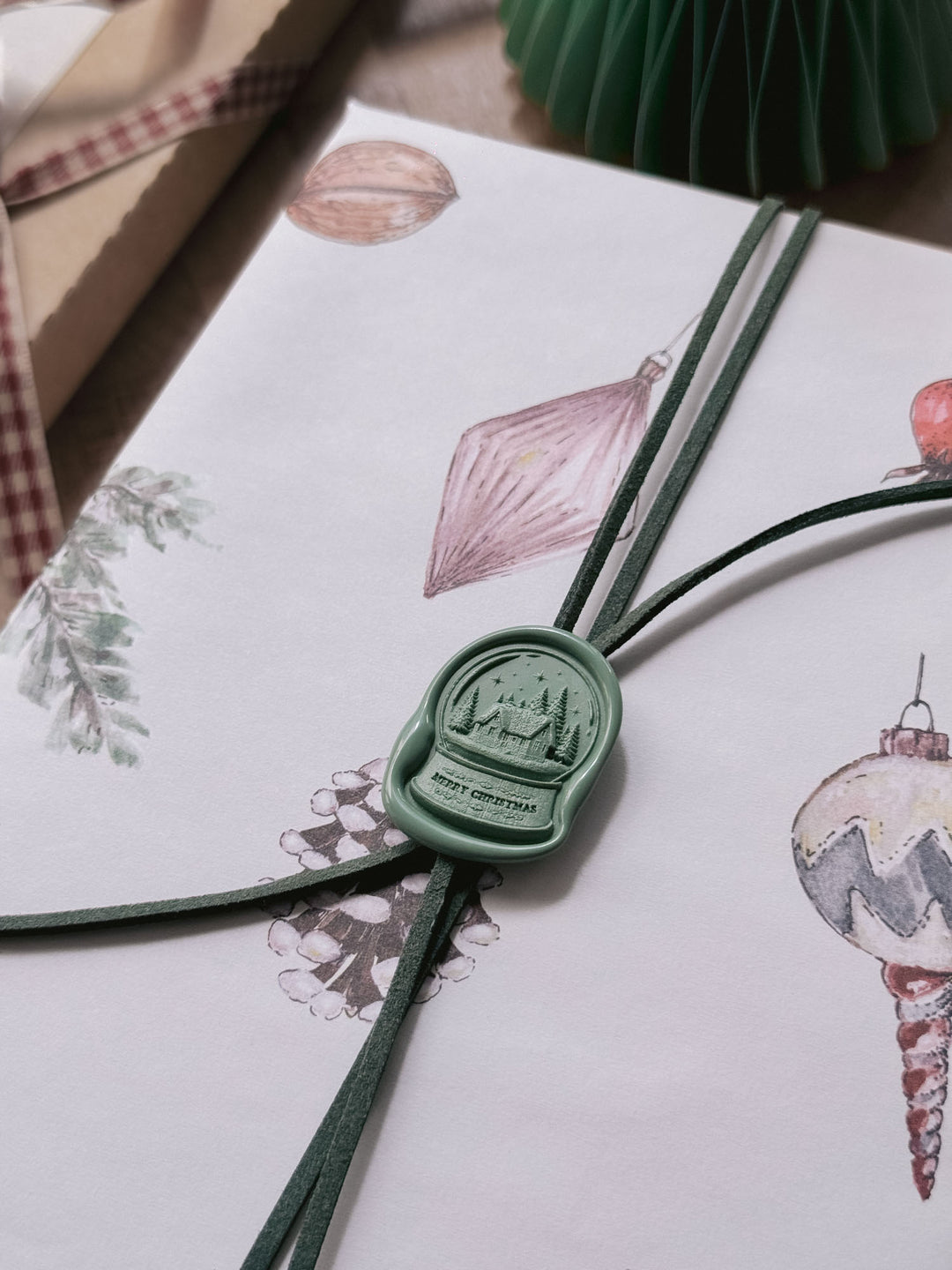 Close-up of a sage green die-cut snow globe wax seal with Merry Christmas lettering, securing green suede cord on illustrated Christmas gift wrap.