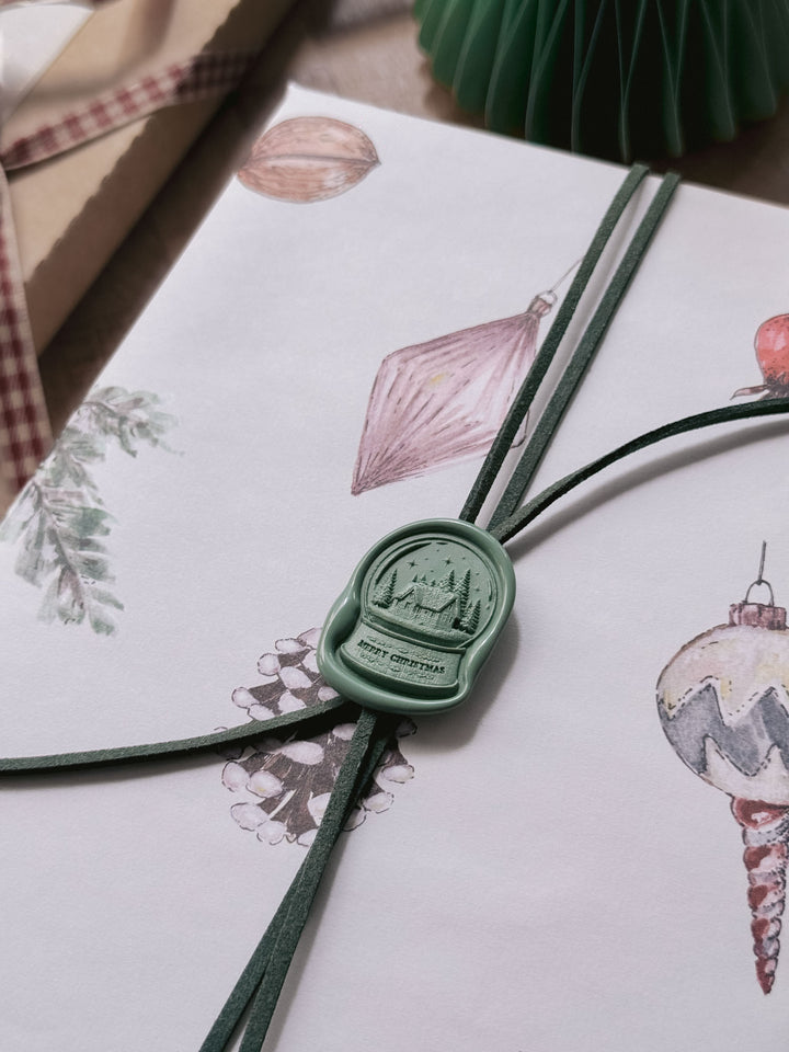 Close-up of a sage green die-cut snow globe wax seal with Merry Christmas lettering, securing green suede cord on illustrated Christmas gift wrap.