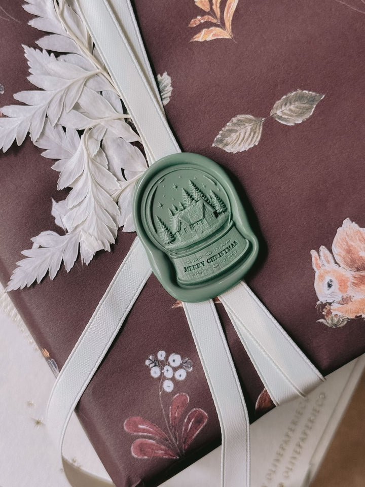 Sage green die-cut Merry Christmas snow globe wax seal on festive burgundy gift wrap, finished with ivory ribbon and dried white foliage.