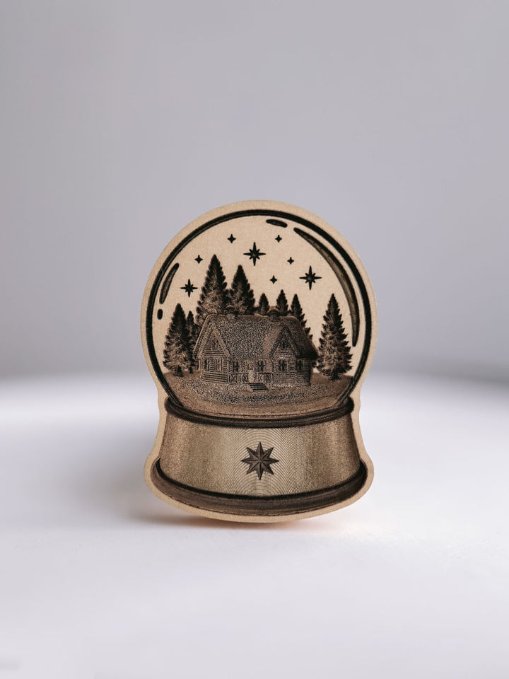 Snow globe wax seal stamp, showing the die-cut brass stamp head engraved with a cabin, pine trees and a north star on the base, against a white background.