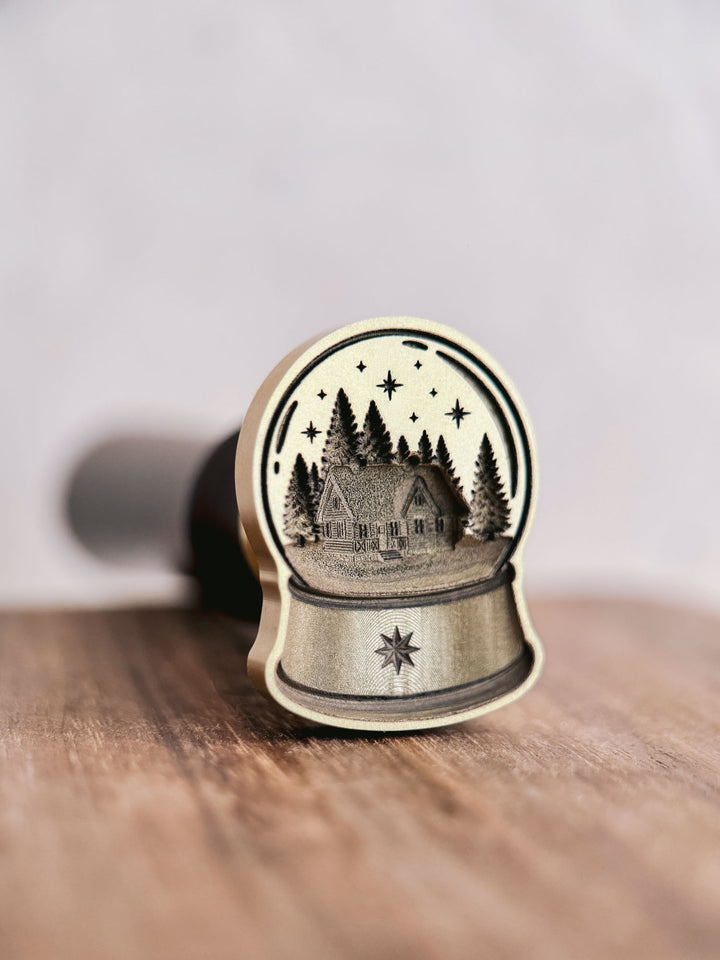 Die-cut brass snow globe wax seal stamp with north star detail, standing upright on a wooden surface with the wooden handle behind.