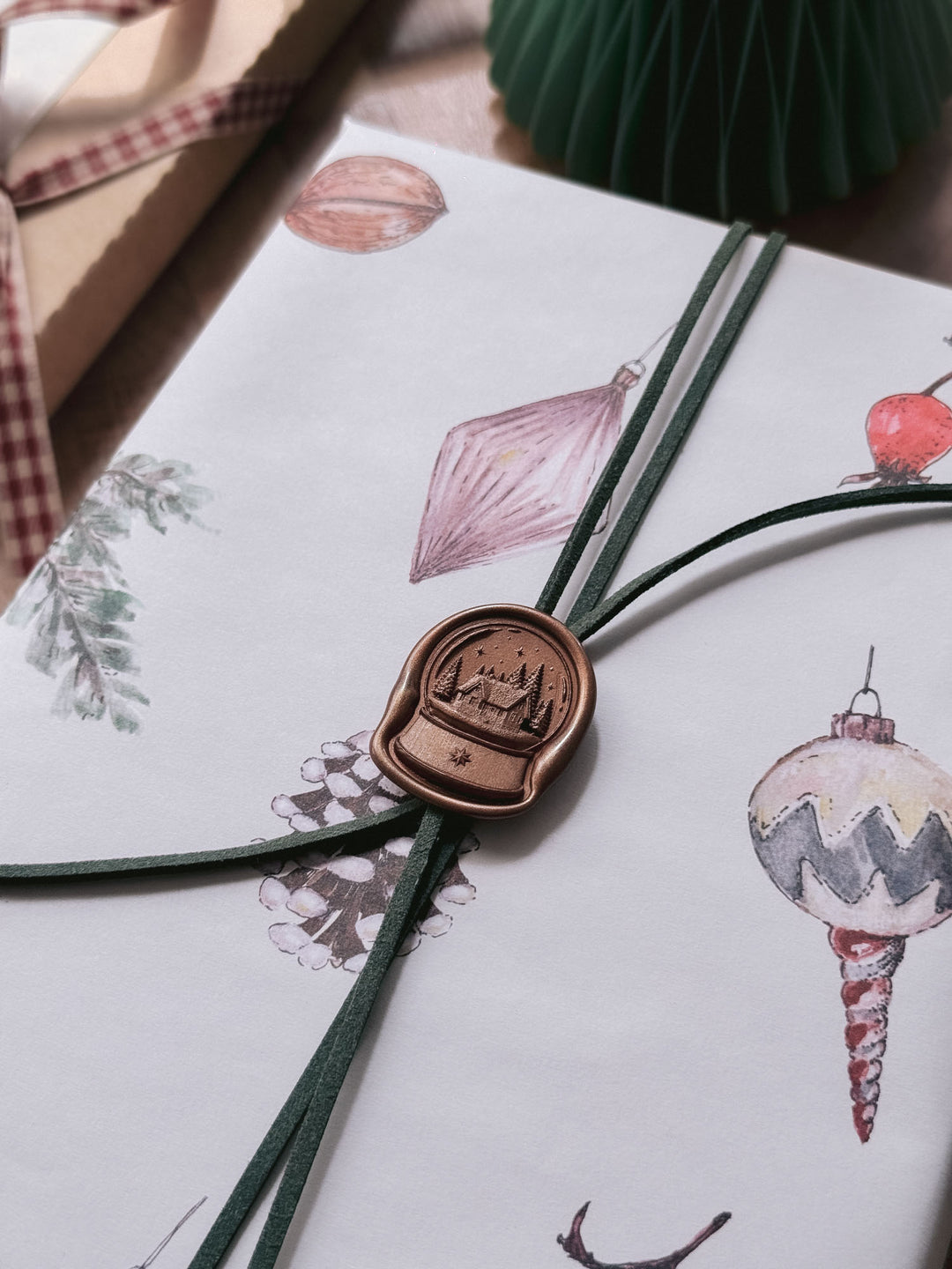 Antique bronze die-cut snow globe wax seal securing green suede cord on festive Christmas ornament gift wrap.