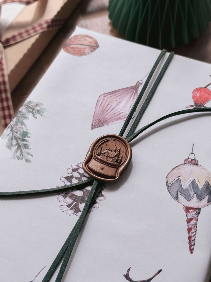 Antique bronze die-cut snow globe wax seal securing green suede cord on festive Christmas ornament gift wrap.