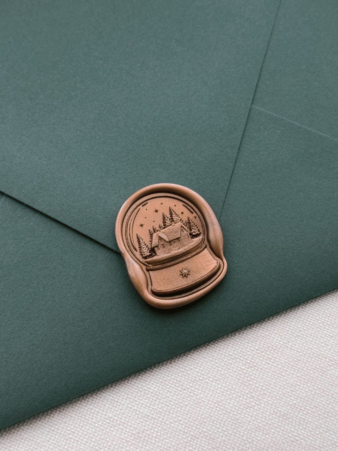 Bronze colored snow globe wax seal featuring a cabin in a forest under a starry sky, on a dark green Christmas card envelope 