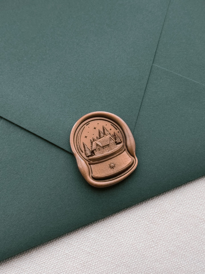 Bronze colored snow globe wax seal featuring a cabin in a forest under a starry sky, on a dark green Christmas card envelope 