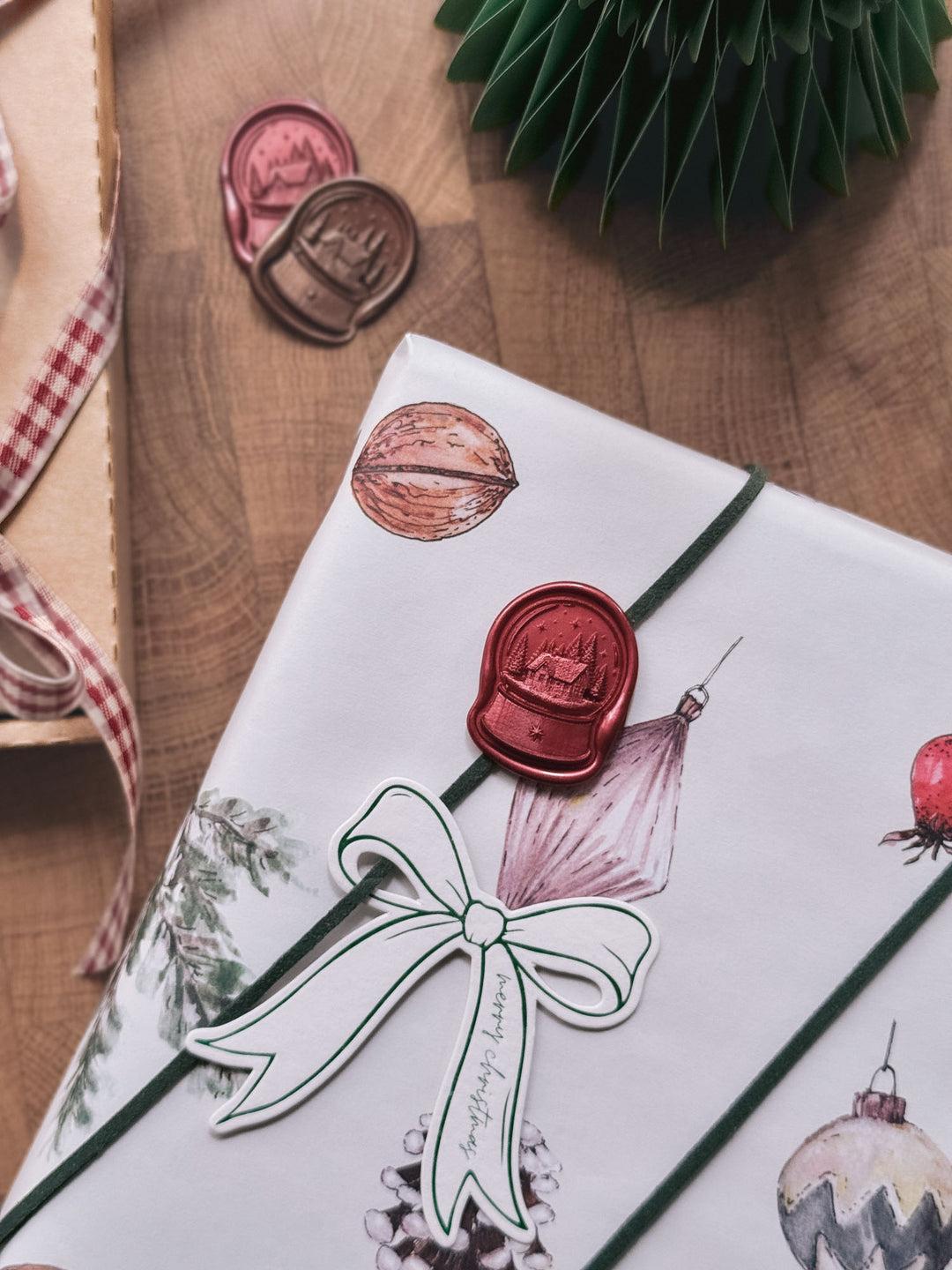 Dark metallic red die-cut snow globe wax seal on white ornament-print gift wrap, with a bow-shaped gift tag and loose wax seals on the wooden surface behind.