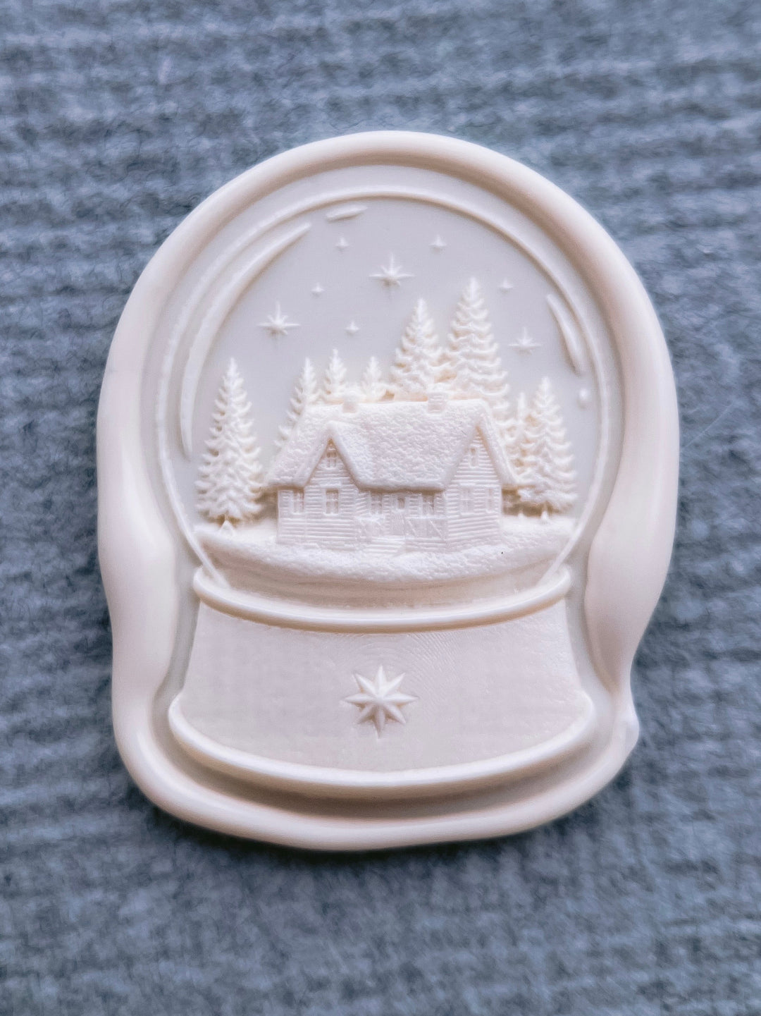 close up shot of an off-white snow globe wax seal in a die-cut shape featuring a snow dusted cabin in a forest under a starry sky, with a 3D North Star engraved at the bottom of the snow globe