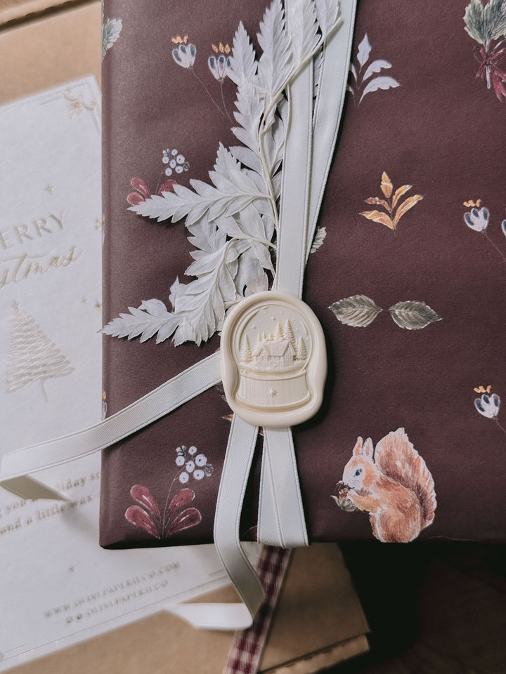 Off-white die-cut snow globe wax seal on festive burgundy woodland gift wrap, tied with ivory ribbon and dried foliage.