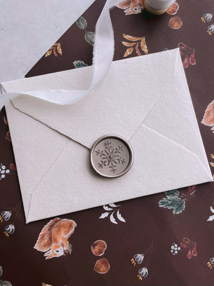 Light bronze 3D engraved snowflake wax seal on a light beige deckle edge envelope on a brown festive background