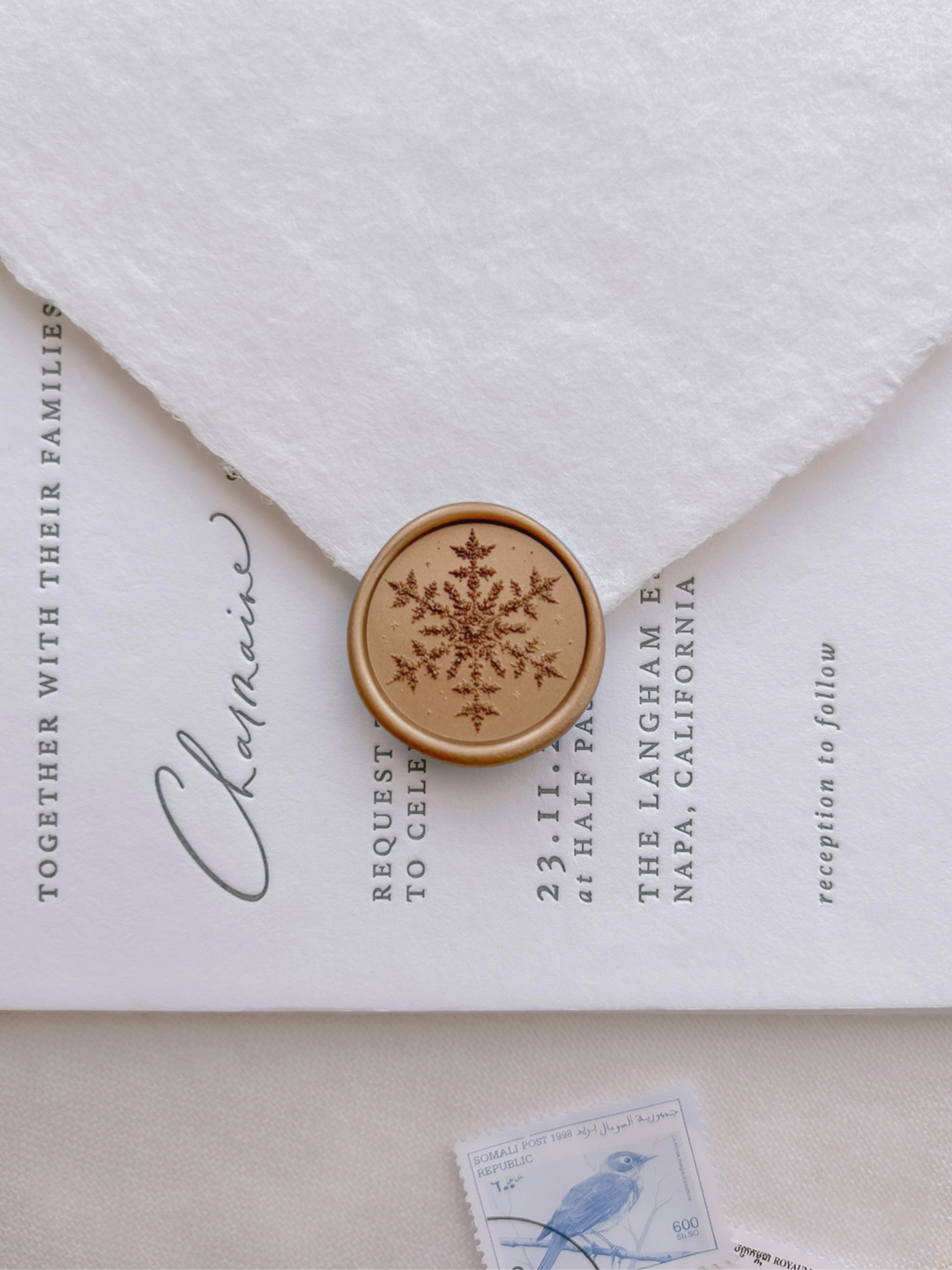 Gold 3D snowflake wax seal on a white deckle edge envelope over a wedding invitation card